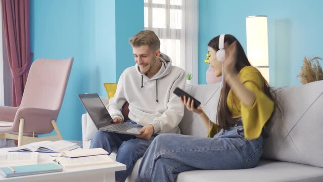 Happy college students having fun at home, laughing, taking a class break. Two fun friends are happy at home, young girl sitting on sofa listening to music young man looking at funny videos on laptop.