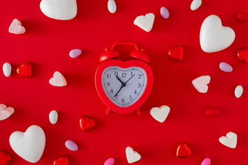 Valentine's Day mood concept. Flat lay photo of heart shaped alarm clock, confectionery chocolate candies and marshmallow on red background. Love Valentines card idea.