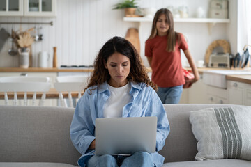 Young woman mother sitting on sofa at home working on laptop while her daughter standing behind with gift box, kid child holding present making surprising mom on her birthday. Selective focus