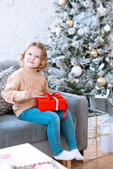 cute happy little girl with curls in Santa hat is sitting on chair near Christmas tree with red gift box in her hands, New Year and Christmas family concept