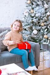 cute happy little girl with curls in Santa hat is sitting on chair near Christmas tree with red gift box in her hands, New Year and Christmas family concept