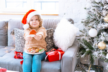 Obraz premium cute happy little girl with curls in Santa hat is sitting on chair near Christmas tree with plate of Christmas gingerbread cookies in her hands, New Year and Christmas family concept