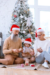 Fototapeta premium Christmas family grandmother, grandfather and granddaughter sit on sofa near Christmas tree and open advent calendar, pretty girl and active happy seniors give each other gift box