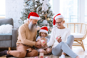 Obraz premium Christmas family grandmother, grandfather and granddaughter sit on sofa near Christmas tree and open advent calendar, pretty girl and active happy seniors give each other gift box