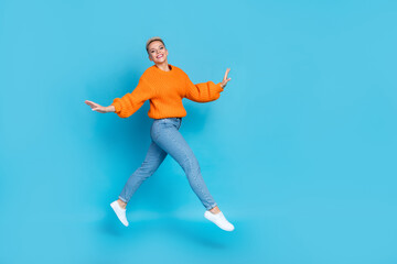 Fototapeta na wymiar Full length photo of excited carefree lady wear orange sweatshirt jumping high walking empty space isolated blue color background