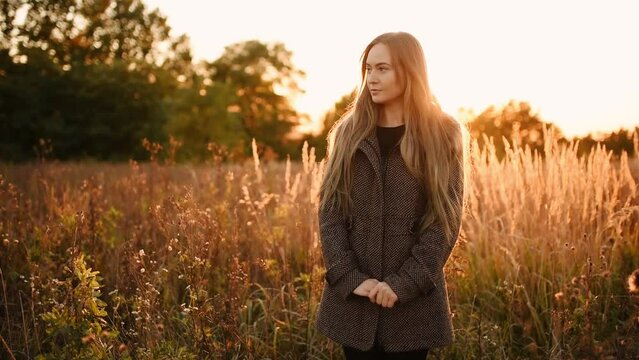 fair-haired long blonde young girl is standing field in woolen short coat at sunset. beautiful sunset rays of sun break through high dry stalks of grass. high-quality video. woman feel cold outside