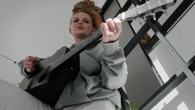 woman with dreadlocks on head singing song and playing acoustic guitar in sunny cottage bedroom, simple living and hobby relaxing