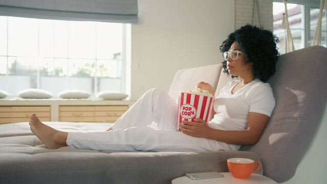 Attractive woman sitting on couch sofa with popcorn basket at home. African American female watching entertaining movie on TV. Female really scared by thriller movie plot scenario and special effects