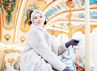 Obraz na płótnie Canvas Beautiful and cheerful brunette woman in a fur coat rides on a carousel