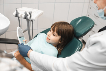 Medicine, stomatology, technology and health care concept. Little girl looking in the mirror after dental procedure.