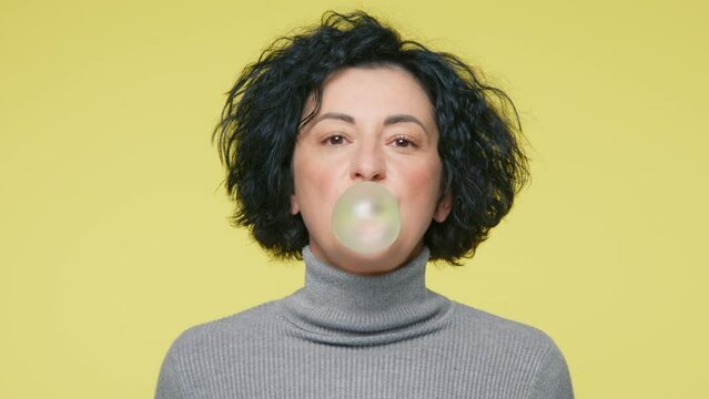 Smiling mature woman inflates ball bursts it isolated on yellow studio wall background. People lifestyle concept 40s years old in grey sweater posing looking camera chewing blowing bubble gum balloon