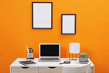 Workplace with laptop, stationery and magnifying glass on desk near orange wall. Home office