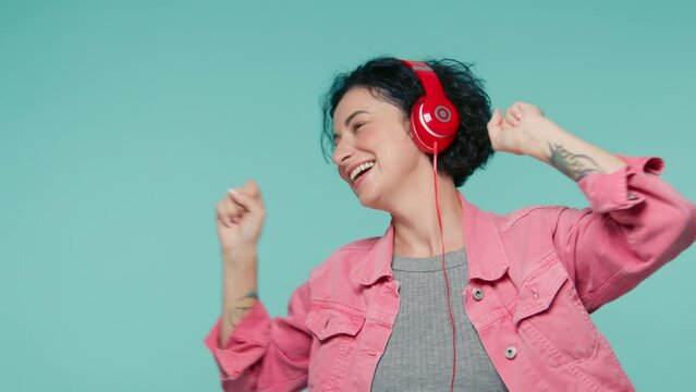 Slow motion sliming female in pink denim jacket and in red headphones dancing indoors, listening music with closed eyes, having fun alone. Happy mature woman 40s dancing in studio on blue background