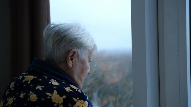 Depressed senior woman at home feeling sad. Elderly woman looks sadly outside the window. Depressed lonely lady standing alone and looking through the window