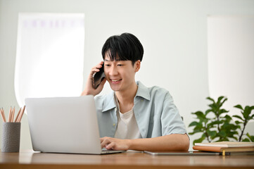 Handsome Asian man talking on the phone and using his laptop computer. work from home