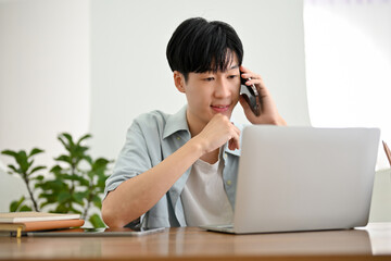 Handsome Asian man on the phone with someone while looking something at his laptop screen