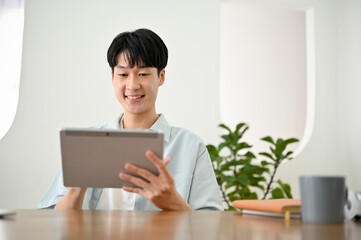 Happy cute young Asian man using his digital tablet at his desk