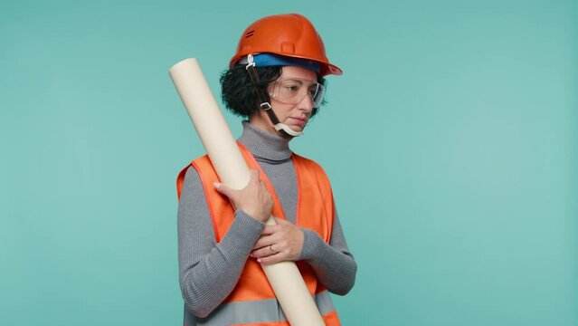 Mature caucasian female civil engineer in bright orange safety jacket and helmet holding many blueprints of the structure, holding it on blue background. Under construction building working together