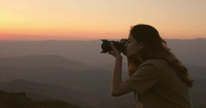 A woman photographer against the background of a foggy hilly valley takes beautiful pictures of an orange-pink sunset. A woman captures the landscape of the setting sun in the mountains with a camera.