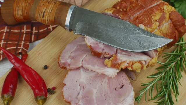 Sliced slices of juicy pork ham on wooden cutting board, next to it are a carving knife, multi-colored allspice peas, red hot peppers, parsley, dill and basil. The concept of delicious meat products.