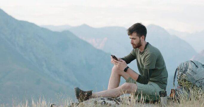 A happy young hipster backpacker is sitting on top of a mountain and surfing the internet on his phone. A male resting on a rock watches the sunset, looks into the distance and dreams.