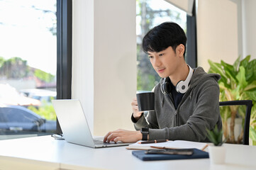 Smart young Asian male programmer or web developer sipping morning coffee while using laptop