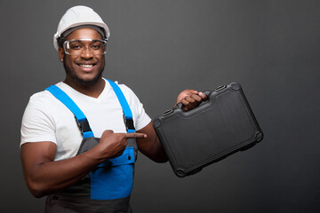 Cheerful African American man in robe and protective helmet demonstrates box with tools for auto...
