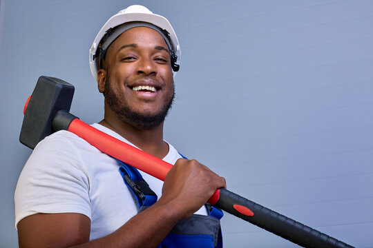 Big Black Guy In A Protective Helmet And Work Clothes Looks At The Camera And Smiles While Holding A Hammer On His Shoulder. Portrait Of An African-American Worker In Overalls