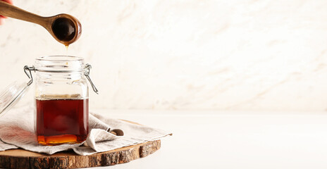 Pouring of maple syrup from spoon into jar on light background with space for text