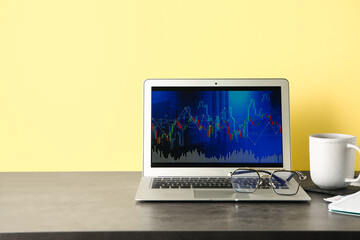 Laptop with stock data and cup of coffee on table near yellow wall. Finance trading