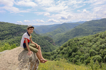 young boy sitting on a stone with beautiful green mountains and blue sky on background