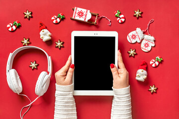 top view female hands holding tablet with black digital screen, Christmas decor, headphones on red table Flat lay Greeting electronic card, book, podcast, music Merry Christmas and Happy New Year - 556391666
