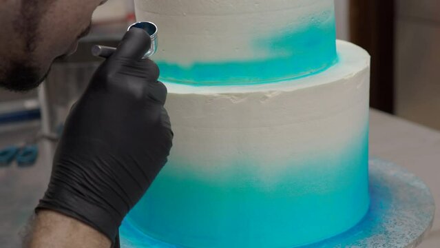 professional baker using edible airbrush colours to decorate two floor layered frosted cake in blue white