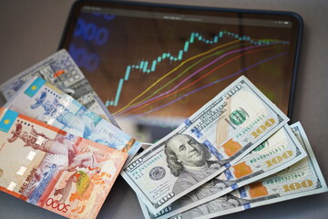 Almaty, Kazakhstan - 10.04.2022 : Stock charts on the tablet and tenge and dollar banknotes on the table.