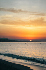 Colorful fiery orange evening sunset on ocean. Silhouettes of surfers in calm sea waves against cloudy sky painted in bright orange. Last rays of sun reflect off surface of the water.