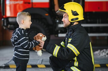 Obraz premium Portrait of rescued little boy with firefighter man standing near fire truck. Firefighter in fire fighting operation.
