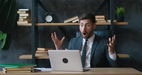 Excited business man looking at laptop screen with enthusiastic wow expression on his face, enjoying lottery win. Happy male entrepreneur in formal suit at modern office celebrating success indoors