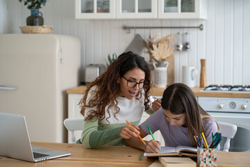 Young mother wearing glasses helping schoolgirl daughter to do homework while sitting together at kitchen table. Teen girl child learning language with tutor at home. Homeschooling concept