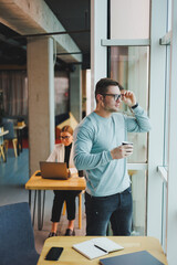 Young promising male manager in casual clothes standing by the window in the workspace and looking at the city. Bright modern office in a skyscraper