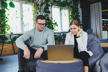 A man and a woman are sitting in a lounge area and using different devices. They are satisfied and...