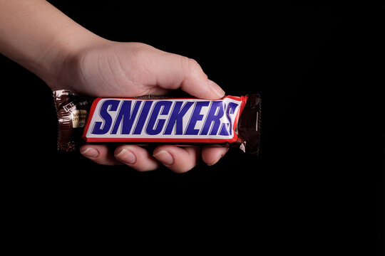 Magdalinovka, Ukraine - December 23, 2022. Female hand holding a snickers chocolate bar on a black background