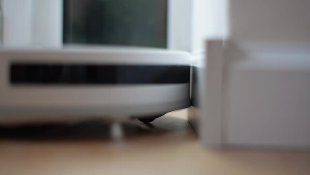 Close-up side view of wireless robot vacuum cleaner vacuuming floor in living room hitting wall and changing direction of movement. Concept of modern smart electronic housekeeping technology.