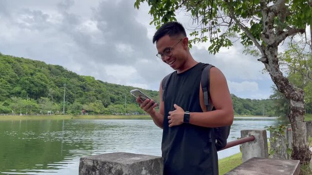Young Filipino man with eyeglasses laughing on video call on cellphone with lake on background. Asian male using phone outdoors, online communication concepts