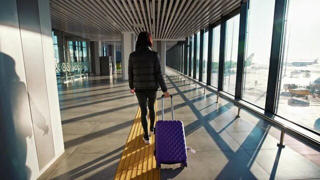 Girl with suitcase walks at airport along corridor with large windows. Back of young business woman passenger walking down walkway. Fly in airplane. Go to rest travel. Vacation and trip, tourism.