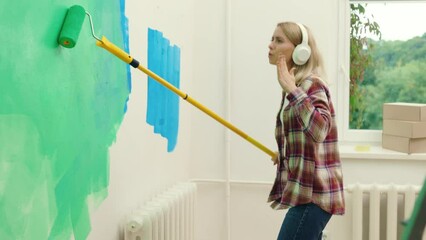 Cheerful woman worker painting wall and dancing enjoying music at home during renovation using earphones. Domestic activities and people concept