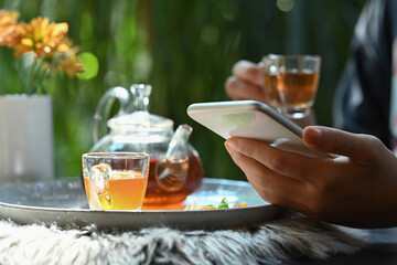 Close up view of woman using smart phone near traditional tea set with cups and teapot on rustic wooden table