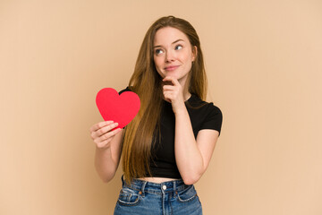 Young redhead woman holding a paper red heart cut out isolated looking sideways with doubtful and skeptical expression.