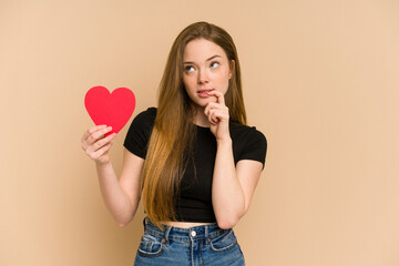 Young redhead woman holding a paper red heart cut out isolated relaxed thinking about something looking at a copy space.