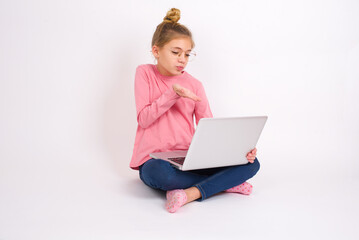 Young beautiful teen girl sitting over white studio background holding a laptop and sending blow kiss to classmates or family via conference call. 