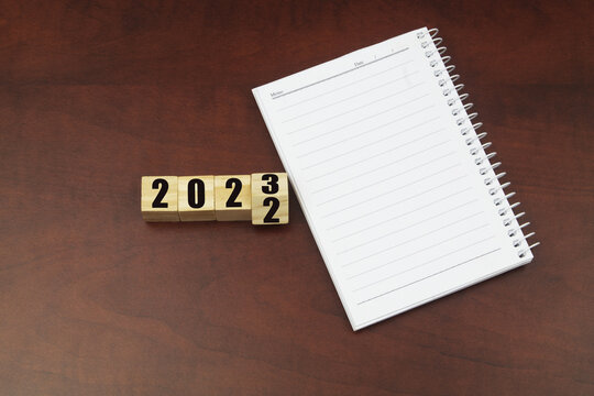 Planning events in 2023 and create goals for new year concept. Numbers 2022 and 2023 on cubes with blank notebook on wooden table. 